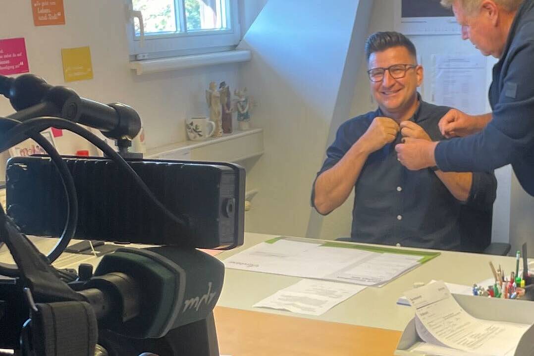 Würdevoll zu Hause: MDR berichtet über die ambulante Palliativversorgung des MVZ Cracau Christian Ptock, Abteilungsleiter der Spezialisierten Ambulanten Palliativversorgung (SAPV), sitzt lächelnd an seinem Schreibtisch und bekommt ein Mikrofon angesteckt. Im Vordergrund ist eine MDR-Kamera zu sehen.